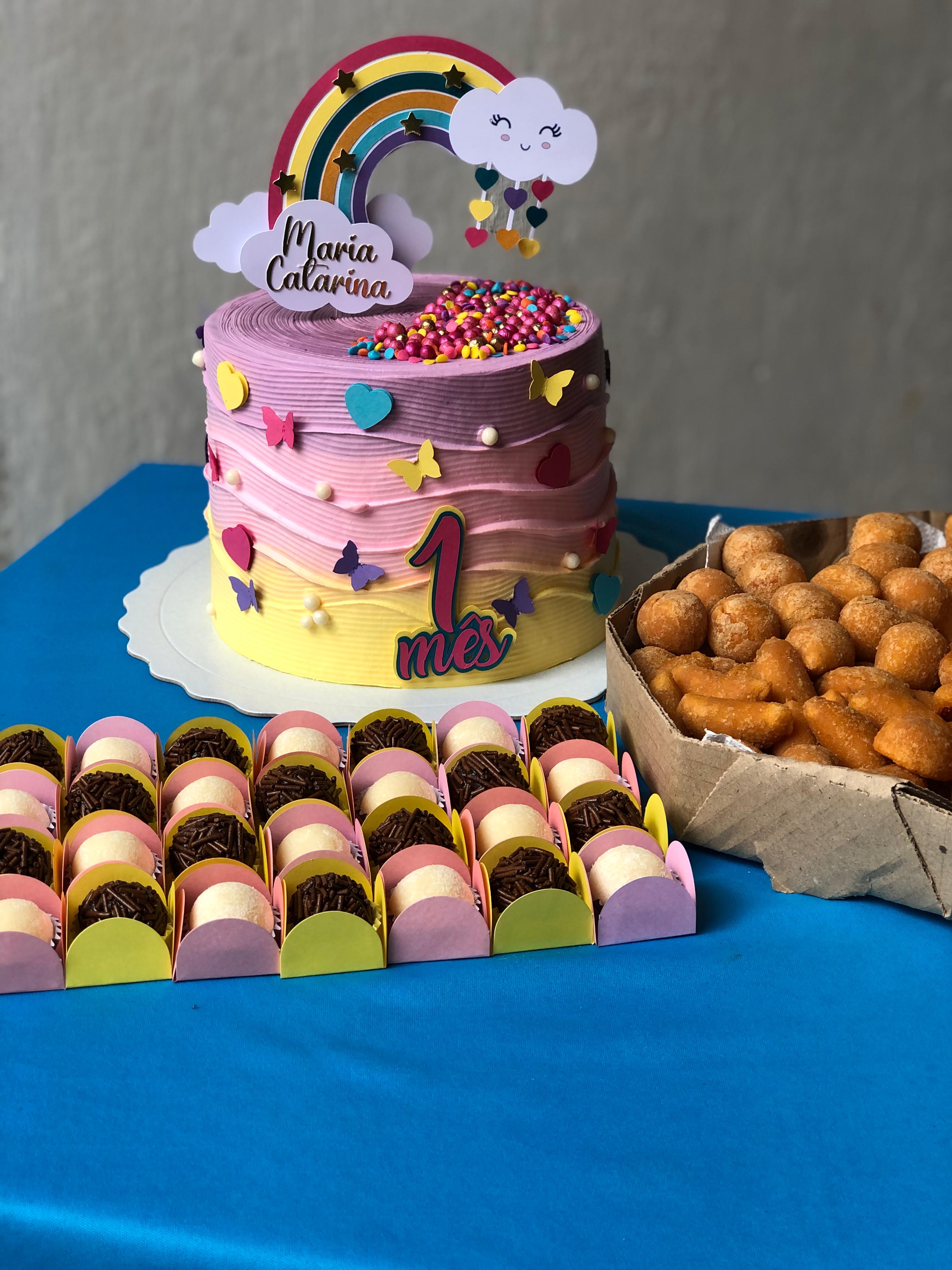 Bolo lilás com arco-íris e decoração colorida, acompanhado de doces e salgados